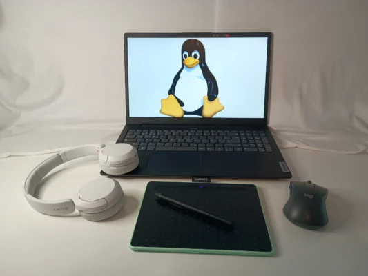 Ein heller Arbeitsplatz zeigt einen Laptop, auf dessen Bildschirm das Tux-Logo zu sehen ist. Links vom Laptop liegen Kopfhörer auf einem Grafiktablett. Eine schwarze Computermaus befindet sich rechts des Tablets.